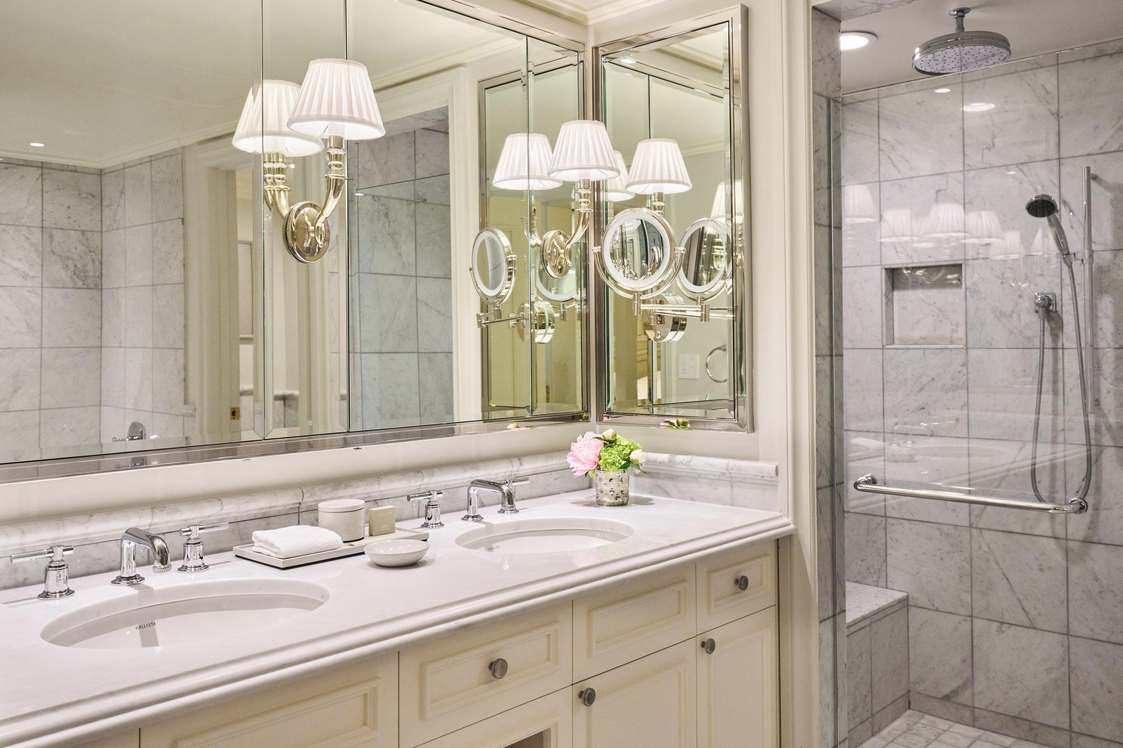 A modern bathroom with dual sinks, a large mirror, wall sconces, and a walk-in shower.