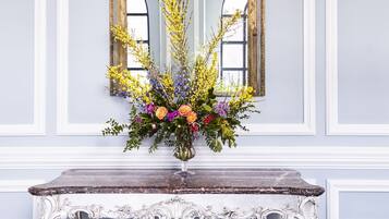 Un espejo con un marco dorado ornamentado, un arreglo floral sobre una consola y un tapete azul estampado.