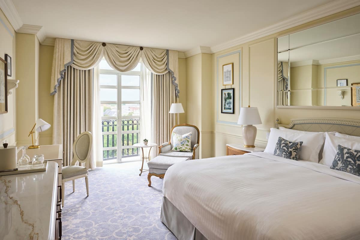 A hotel room with a large bed, a chair, a small table, a lamp, and a window with curtains.