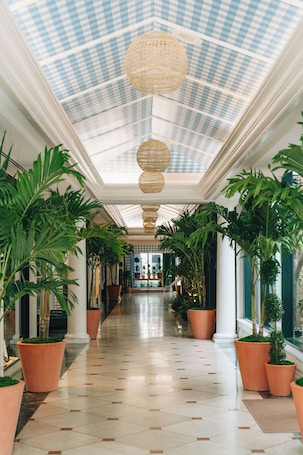 A corridor with a checkered ceiling, large potted plants, and hanging wicker lamps.