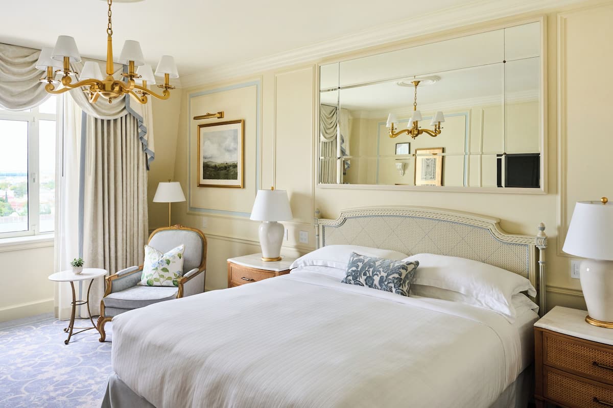 A hotel room with a large bed, a chair, a small table, a lamp, a mirror, and a view of the outdoors.