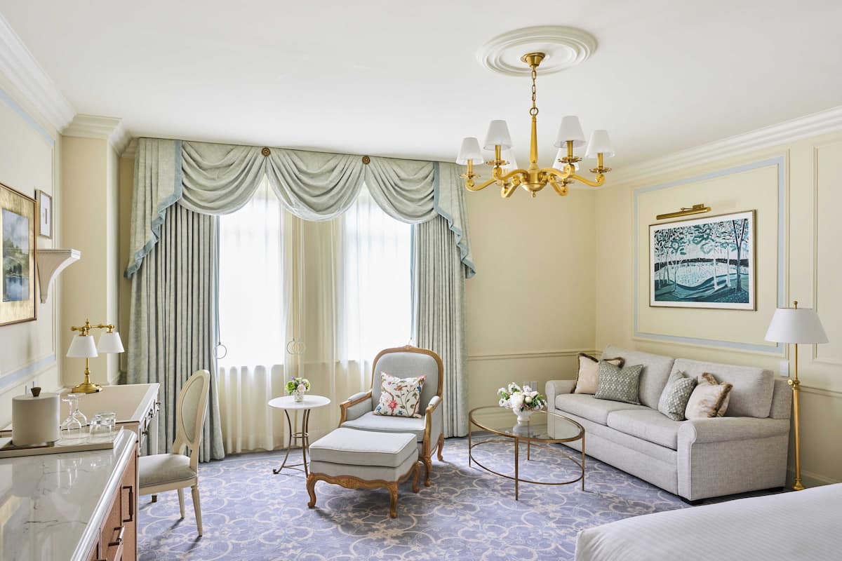 A hotel room with a large window, a sofa, a chair, a small table, and a bed.