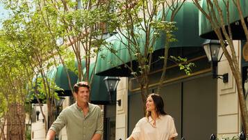 Two people riding bicycles with baskets, a building with green awnings, and a tree-lined street.