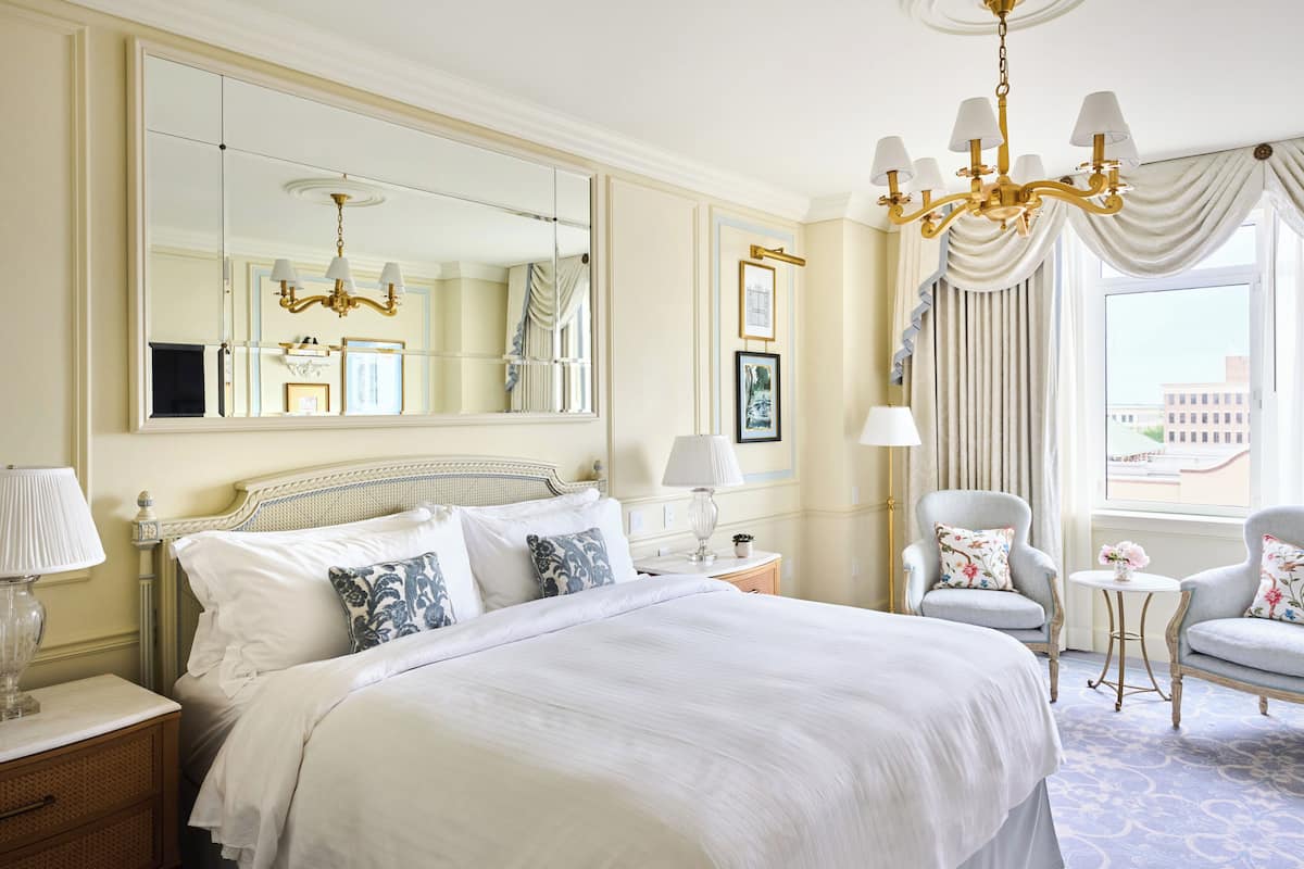 A hotel room with a large bed, a chandelier, a sitting area with two chairs, and a large window with curtains.