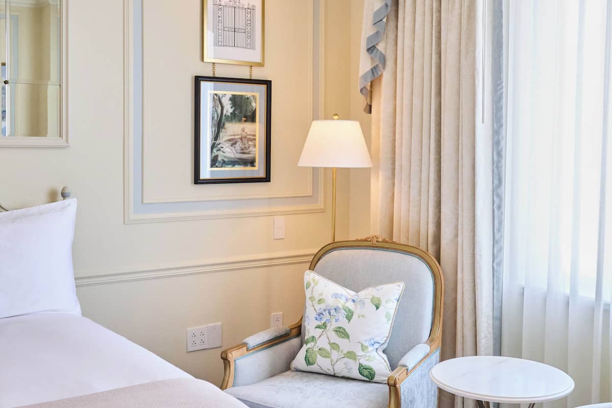A hotel room with a bed, a chair with a floral pillow, a small round table, a lamp, and two framed pictures on the wall.