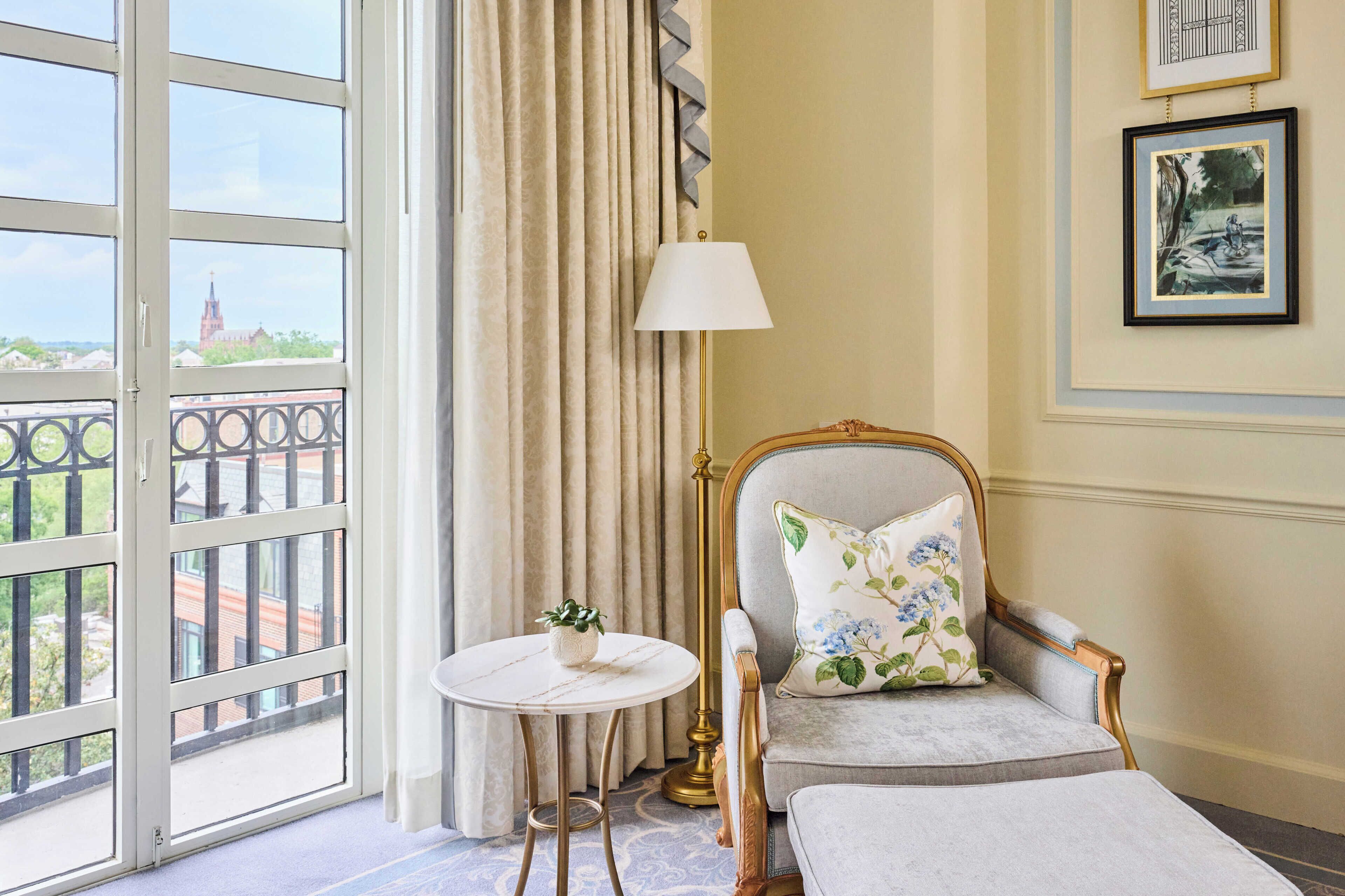 A room with a balcony, a chair with a floral pillow, a small round table with a plant, and a lamp.
