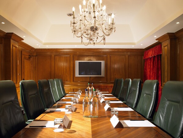 Un salón de conferencias con una larga mesa, sillas de cuero verde, un candelabro y un televisor en la pared.