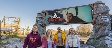 Un grupo de personas caminando por un sendero de madera con una gran pantalla exterior mostrando el texto 'The Bear Grylls Adventure'.