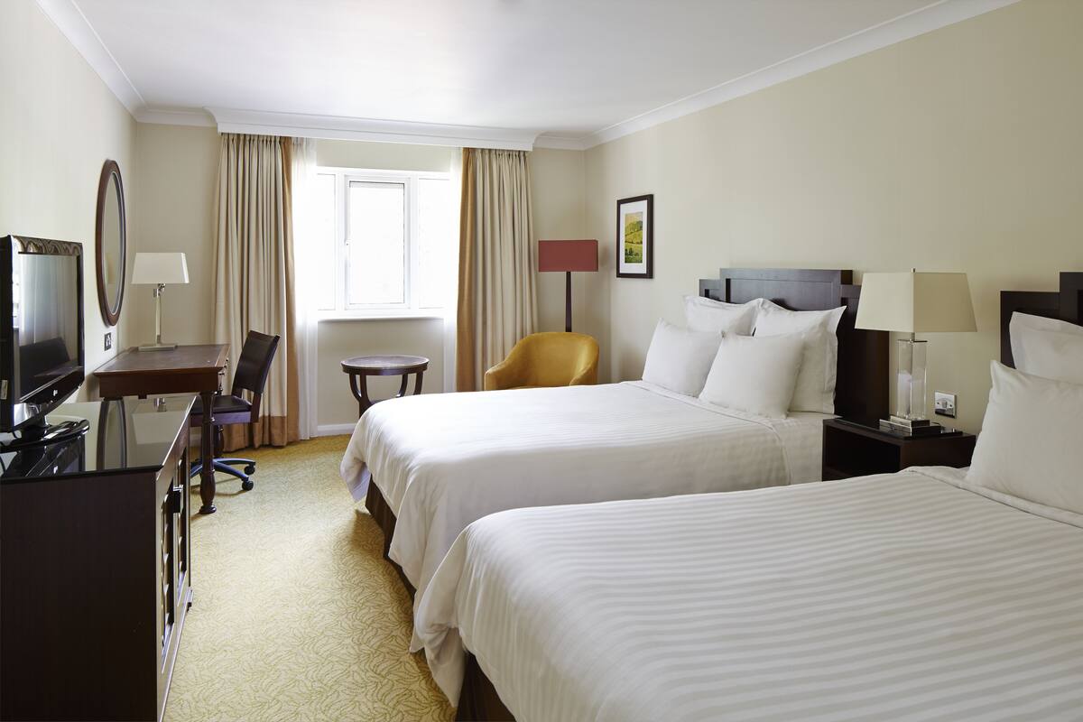 Ein Hotelzimmer mit zwei Betten, einem Schreibtisch mit Fernseher, einem Sessel, einer Lampe und einem Fenster mit Vorhängen.
