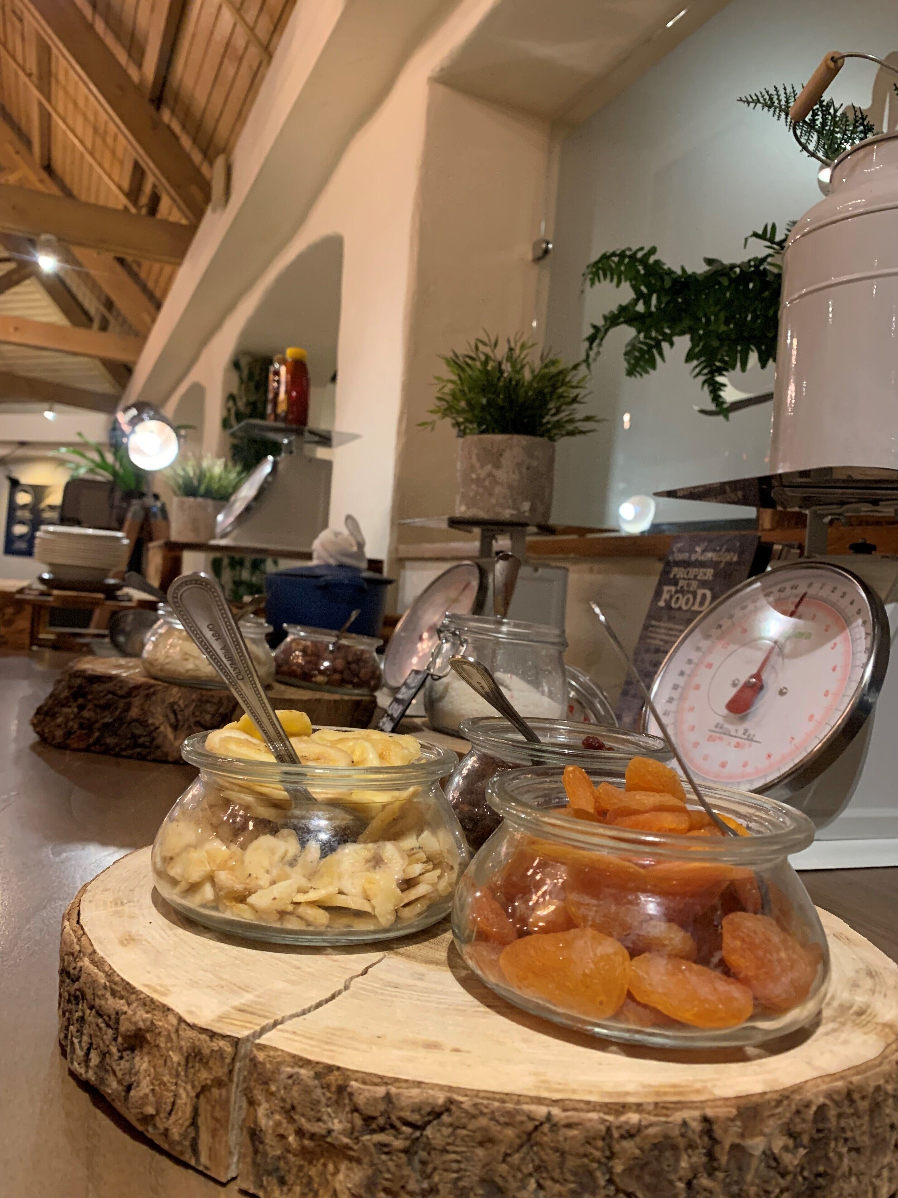 A buffet area with various food items in glass containers on a wooden board, a weighing scale, and a potted plant in the background.