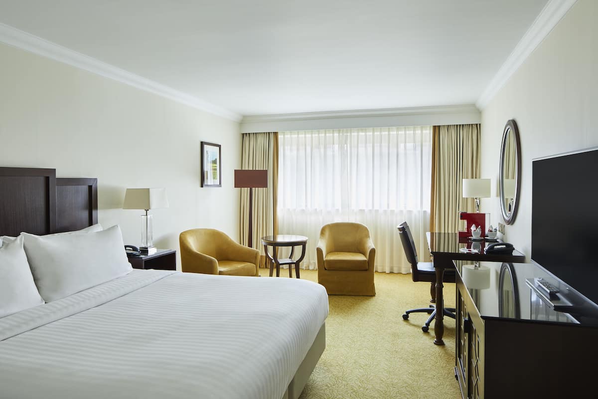 Ein Hotelzimmer mit einem großen Bett, einem Schreibtisch, zwei Sesseln, einem kleinen Tisch, einem Fernseher und einem Fenster mit Vorhängen.
