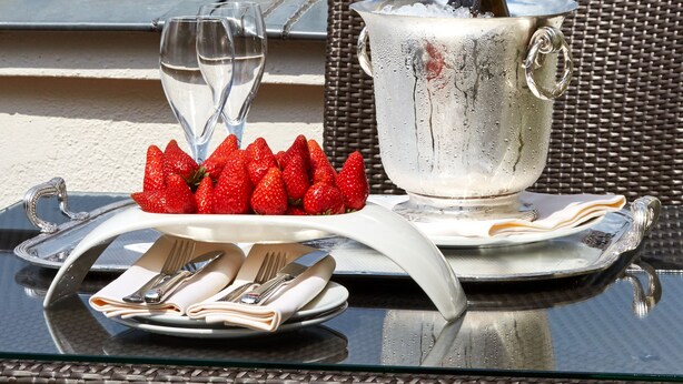 A table with a bottle of champagne, glasses, strawberries, and a plate of cutlery on a balcony overlooking a historic monument.