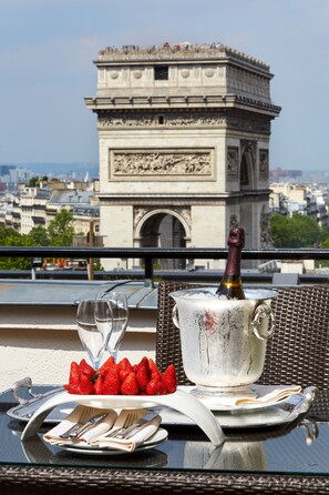 A table with a bottle of champagne, glasses, strawberries, and a plate of cutlery on a balcony overlooking a historic monument.