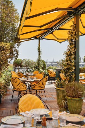 An outdoor dining area with yellow chairs and tables, surrounded by greenery and potted plants.