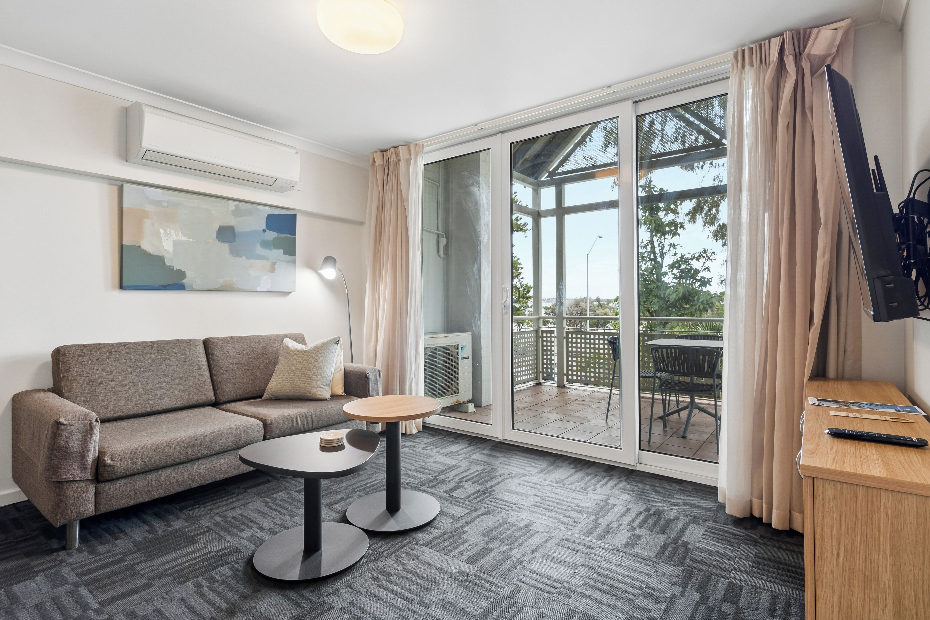 A modern living room with a sofa, a small round table, a flat-screen TV mounted on the wall, and a sliding glass door leading to an outdoor area.