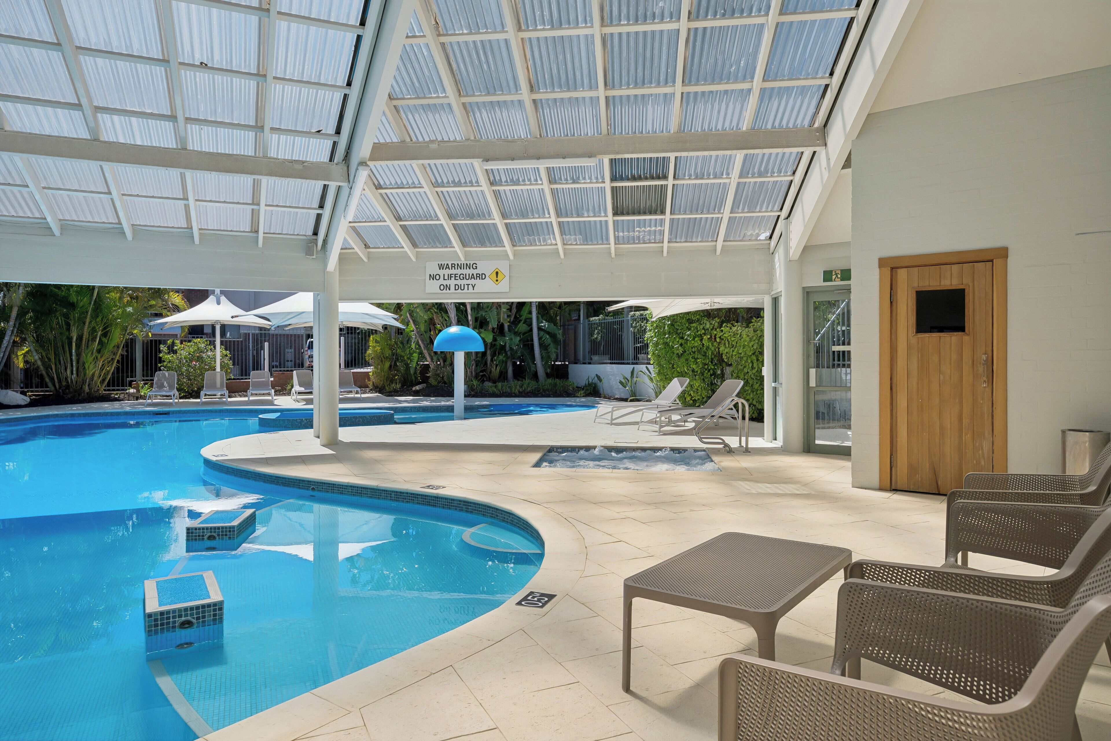 An indoor swimming pool with a glass ceiling, lounge chairs, and a sign indicating no lifeguard on duty.