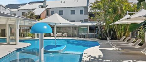 A swimming pool with lounge chairs and umbrellas, surrounded by buildings and greenery.