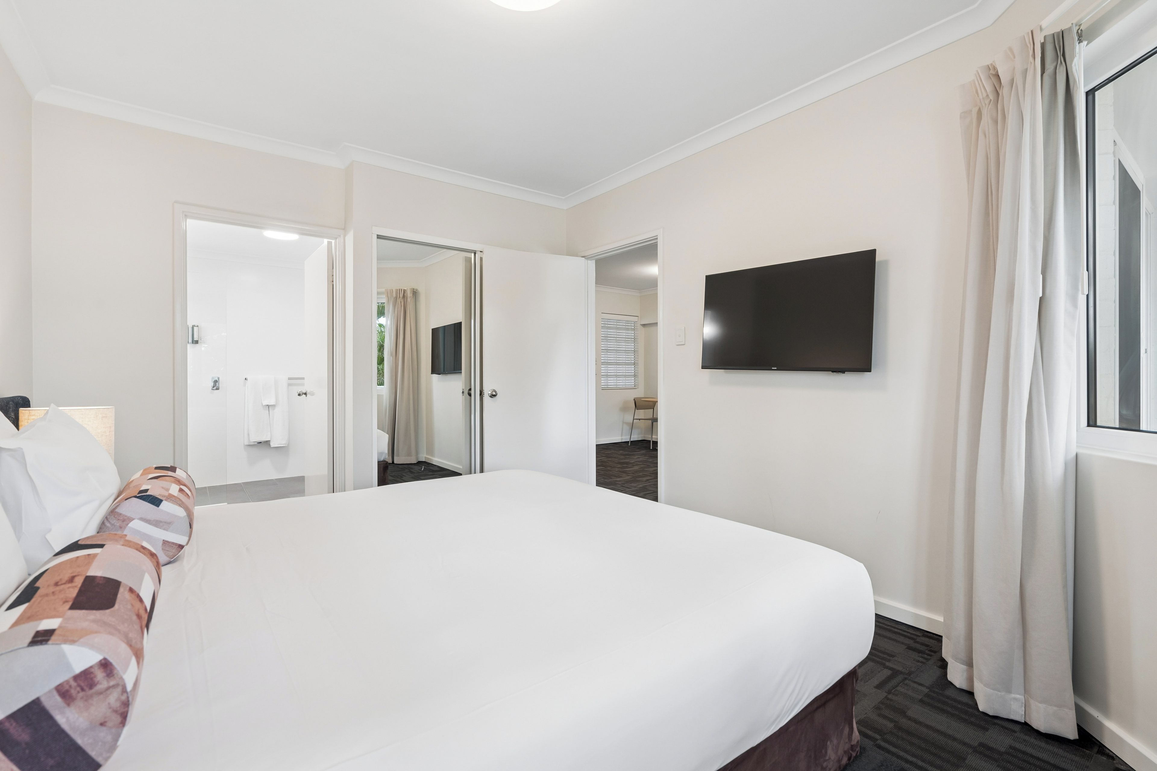A hotel room with a large bed, a flat-screen TV mounted on the wall, and a window with curtains.