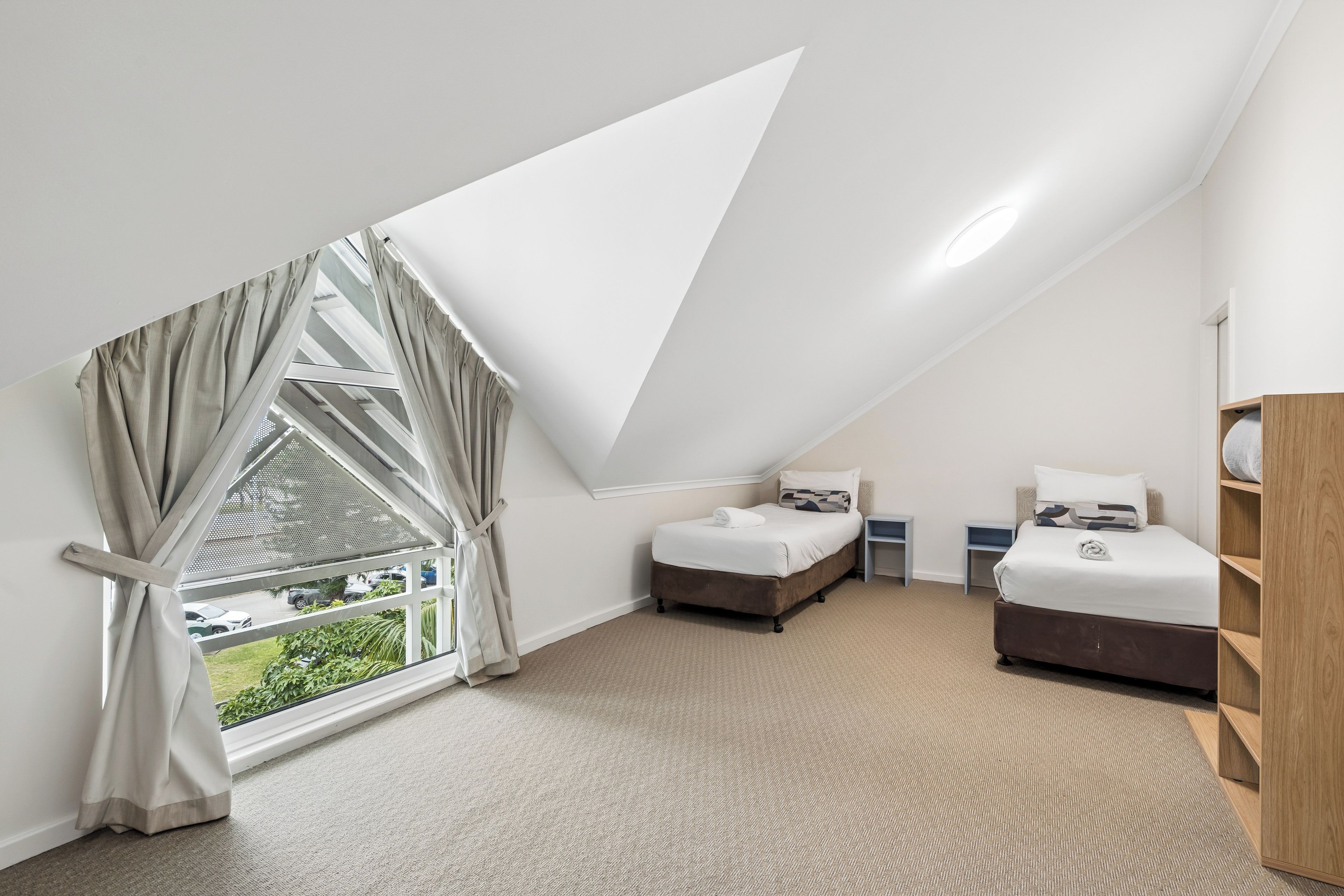 A bedroom with a sloped ceiling, two beds, a wooden wardrobe, and a large window with curtains.