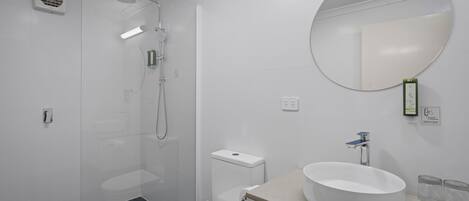 A modern bathroom with a glass-enclosed shower, a white toilet, and a round mirror above a sink with a beige countertop.