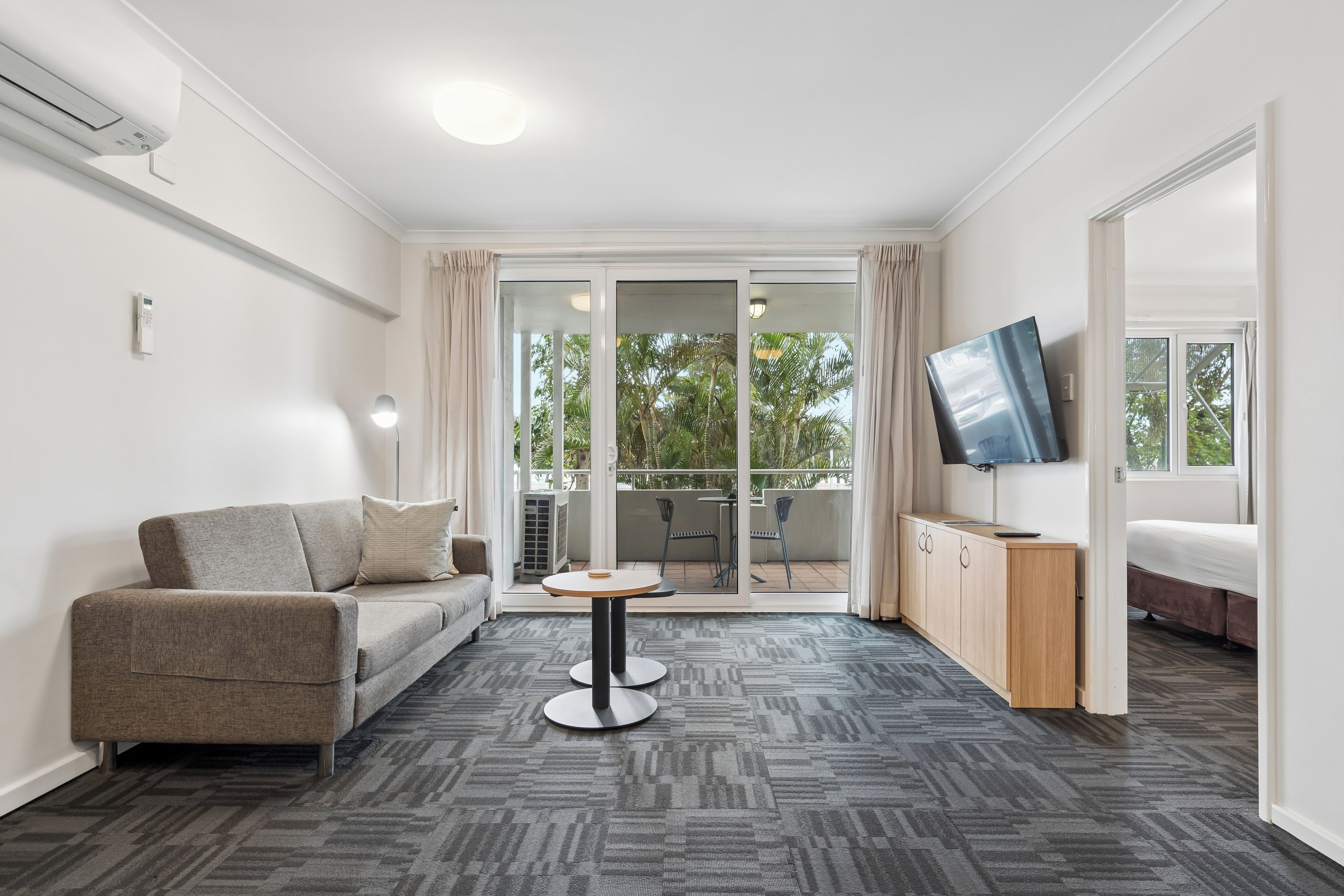 Une chambre d’hôtel moderne équipée d’un canapé, d’une petite table ronde, d’un lit, d’une télévision et d’un balcon avec vue sur les arbres.