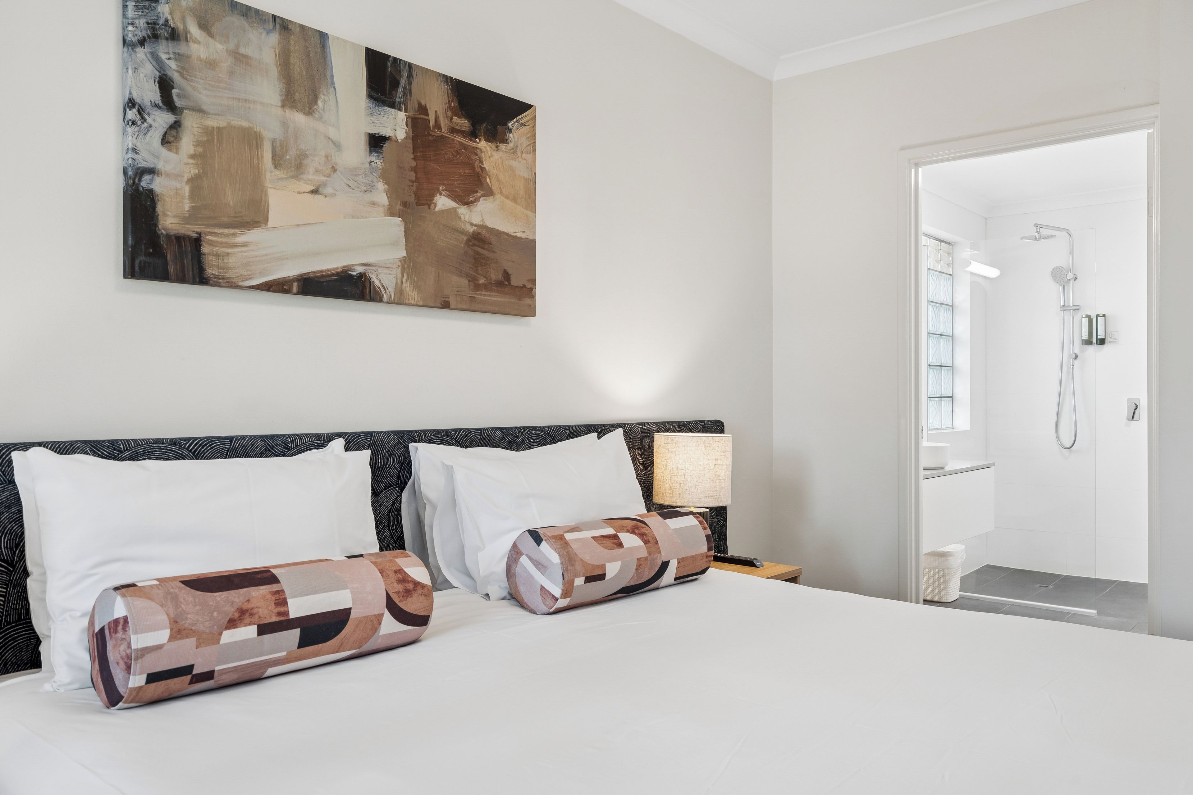 A hotel room with a bed, a nightstand, a lamp, a painting on the wall, and a shower area.