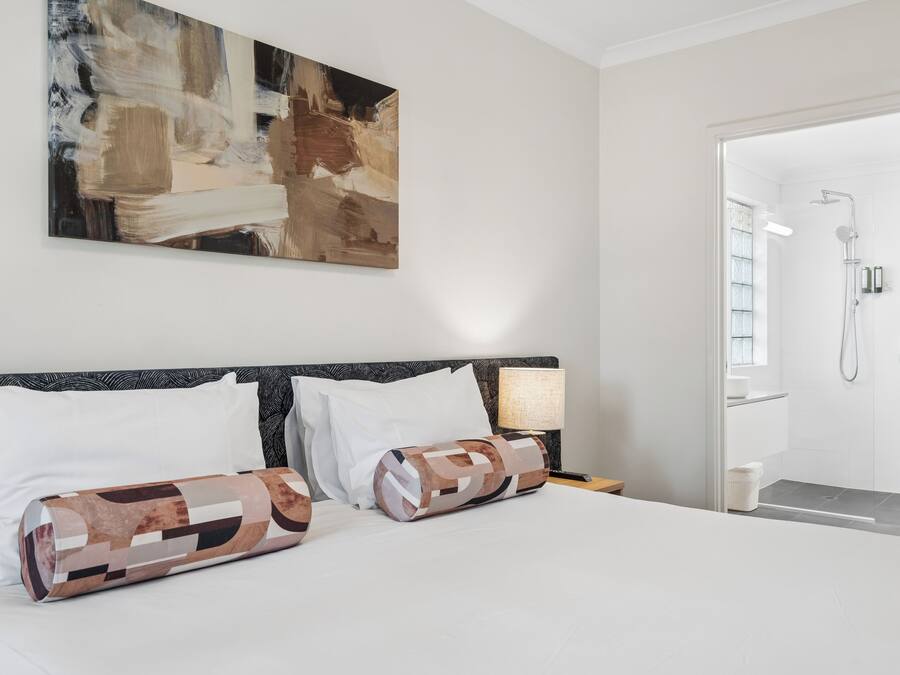 A hotel room with a bed, a nightstand, a lamp, a painting on the wall, and a shower area.