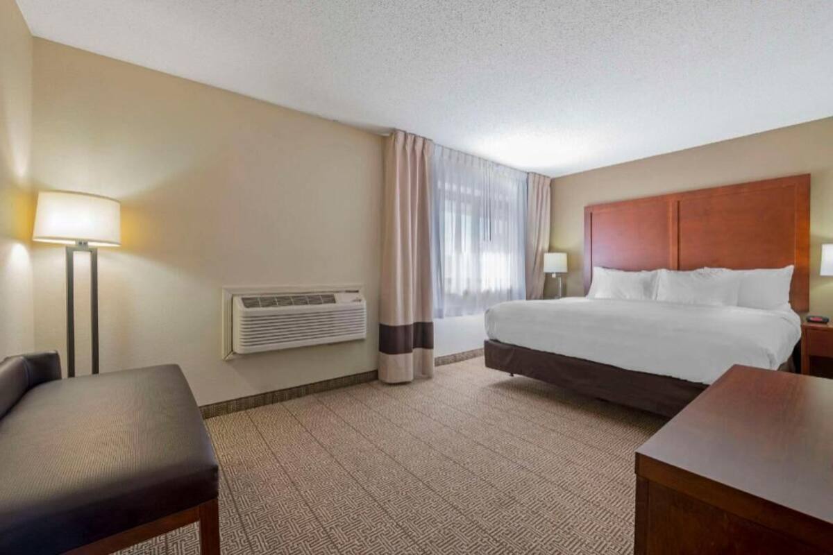 A hotel room with a bed, a desk, a chair, a lamp, and an air conditioning unit.
