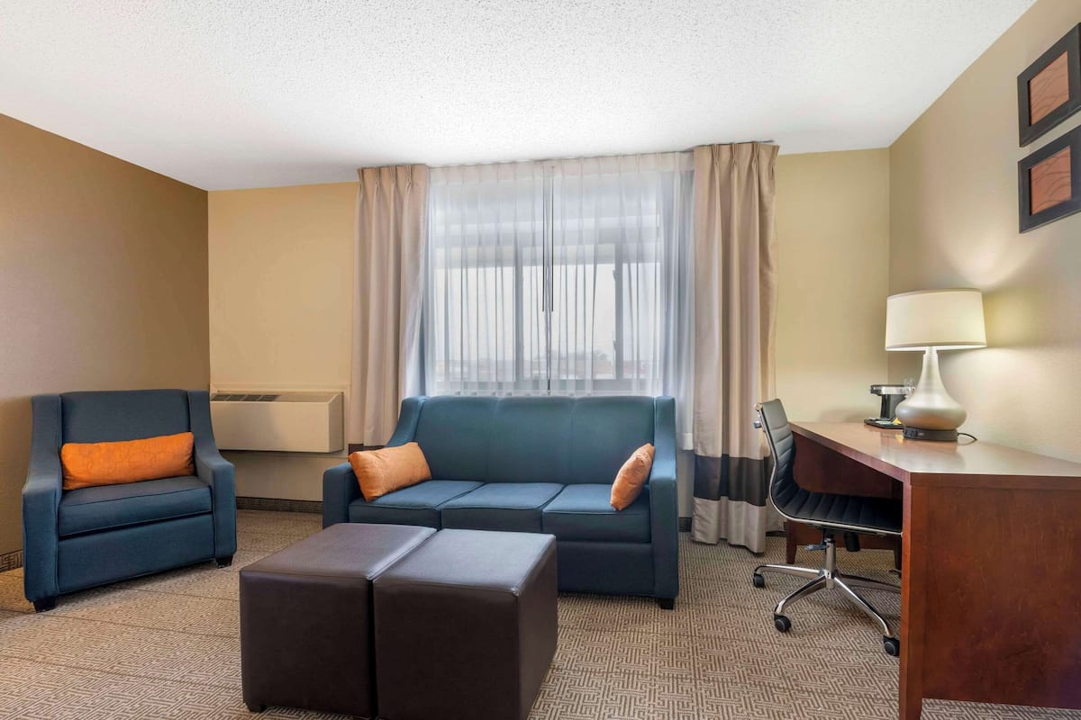 A hotel room with a blue sofa, an orange pillow, a wooden desk, a lamp, and a large window with sheer curtains.