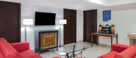 A hotel lobby with red sofas, a glass-top table, a flat-screen TV, a fireplace, and a table with toiletries.