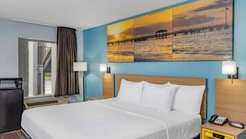 Habitación de hotel con cama, escritorio, lámpara, teléfono y una pintura de un atardecer sobre el agua.