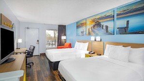 Habitación de hotel con dos camas, un escritorio con televisor, una silla, una lámpara de escritorio y un cuadro de un muelle en la pared.
