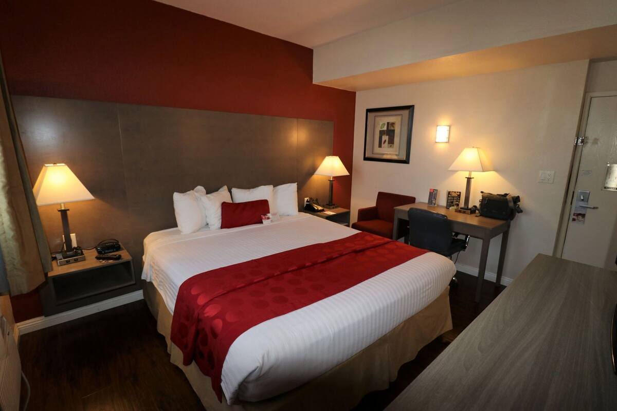 A hotel room with a large bed, two bedside tables with lamps, a desk with a chair, and a door.