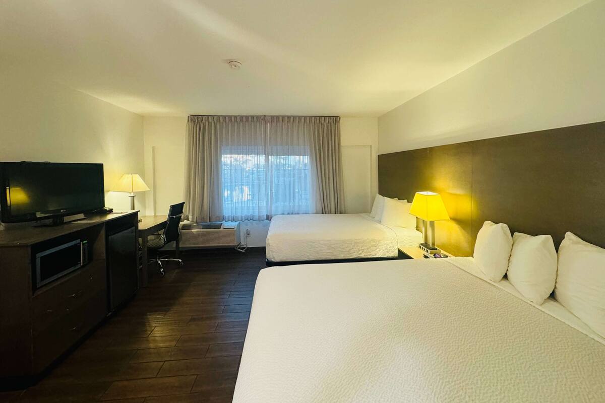 A hotel room with two beds, a TV, a desk with a chair, and a window with curtains.