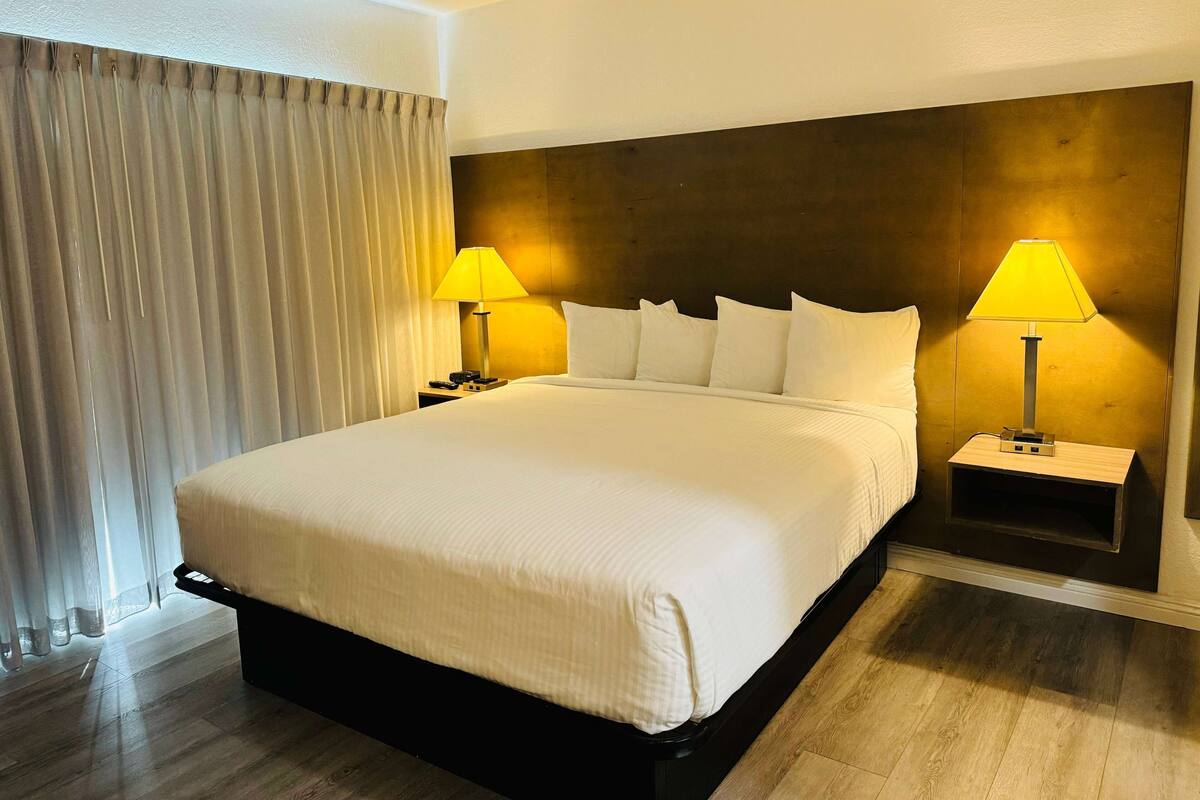 A hotel room with a large bed, two bedside tables with lamps, a wooden headboard, and a curtain on the window.