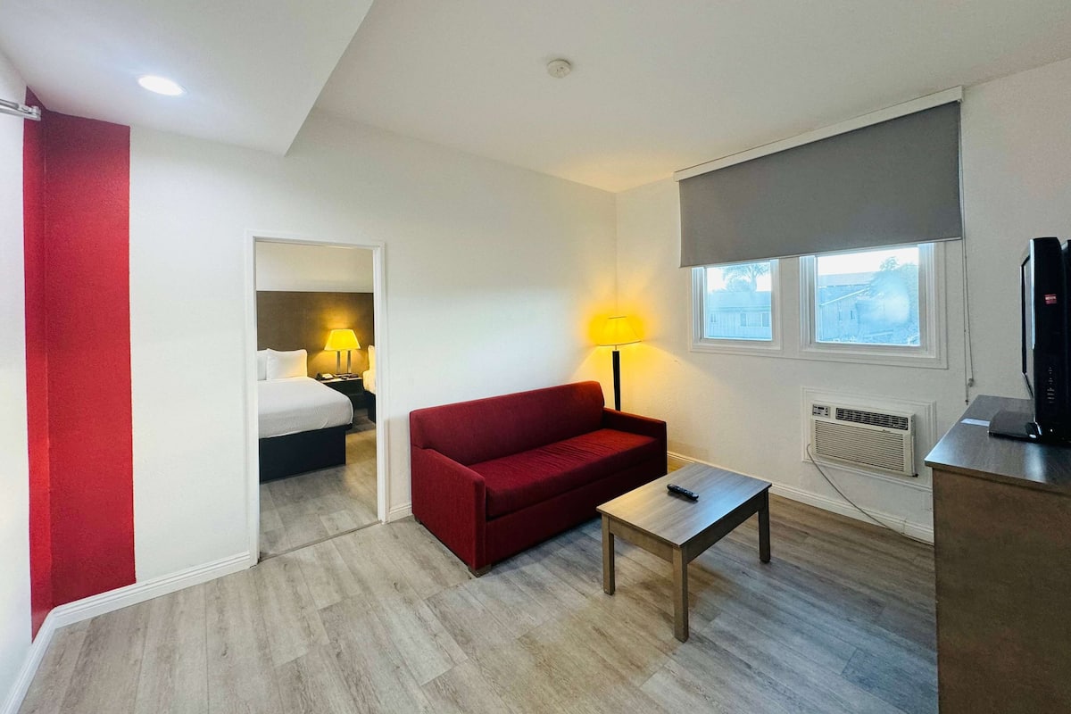 A hotel room with a red sofa, a small table, a lamp, a TV, and a window with a view.