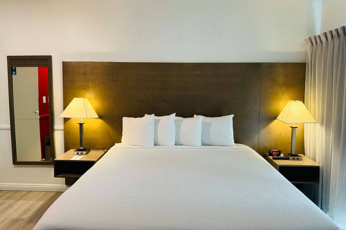 A neatly made bed with white linens, two bedside tables with lamps, a large mirror, and a wooden headboard.