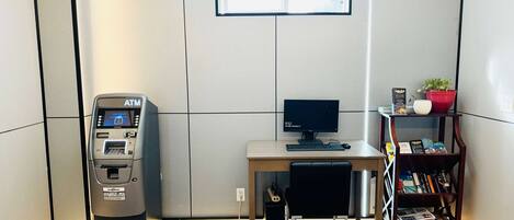 Een kleine, moderne kamer met een bureau, een computer, een geldautomaat en een boekenplank.