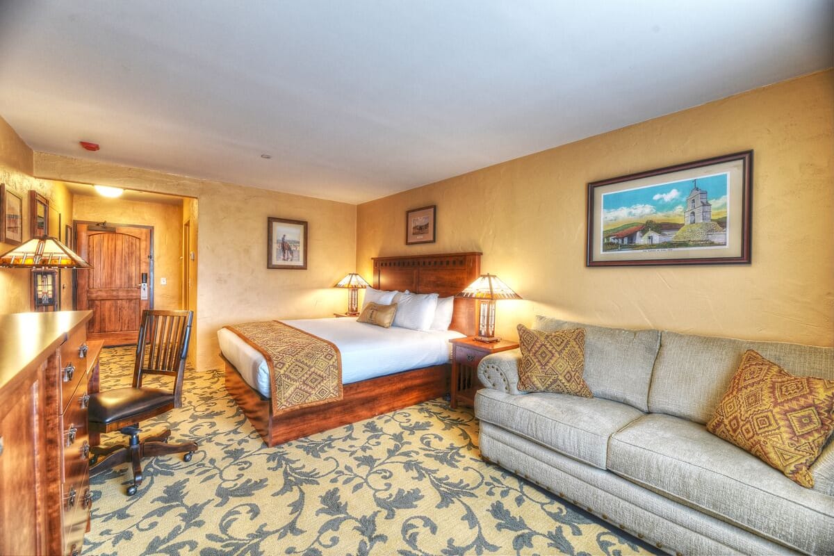 A hotel room with a bed, a sofa, a desk with a chair, and a patterned carpet.