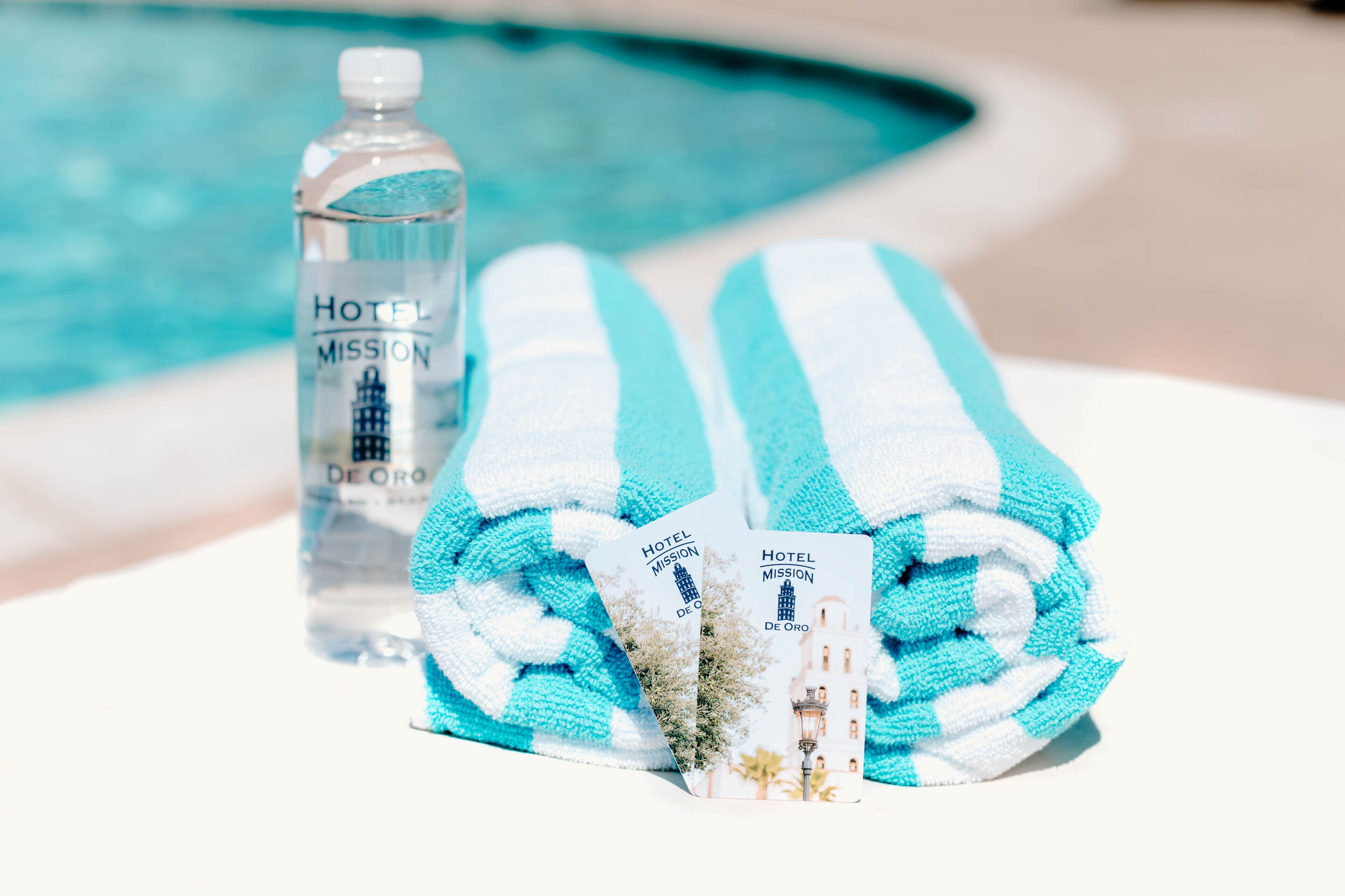 Escena junto a la piscina con una botella de agua etiquetada como "Hotel Mission De Oro" y dos toallas con la misma etiqueta.