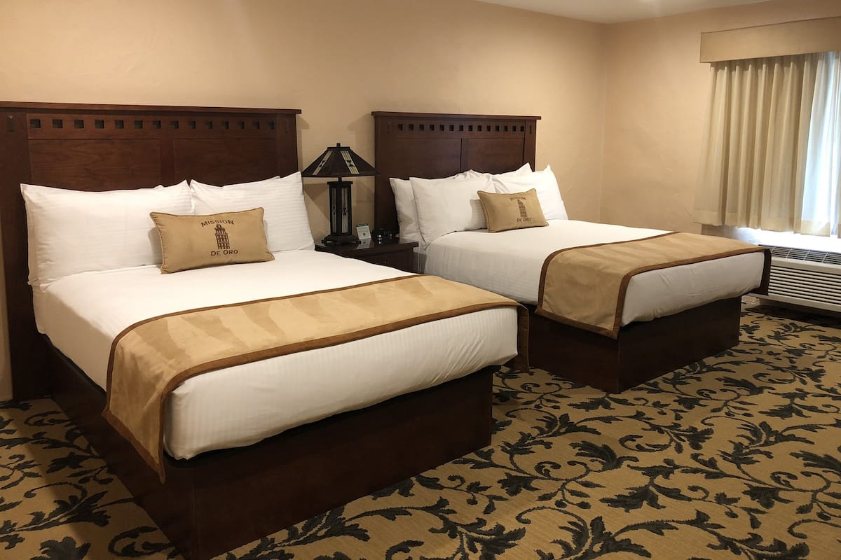 A hotel room with two beds, each with a wooden headboard, white bedding, and beige throw blankets. There is a nightstand with a lamp between the beds, and a window with curtains.