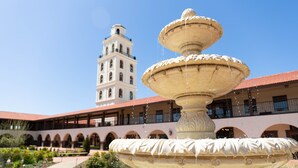 Eine mehrstufige Fontäne in einem Innenhof, im Hintergrund ein Gebäude mit Bogenarkaden und einem Glockenturm.