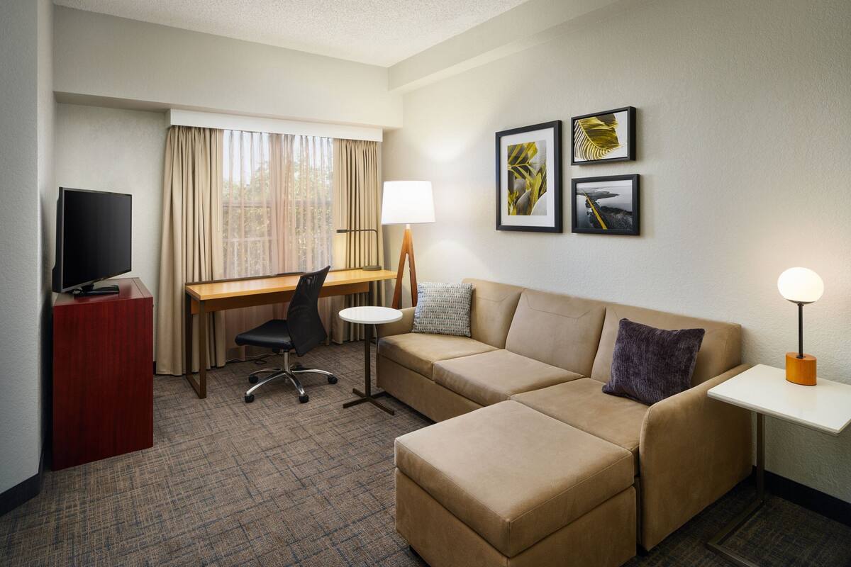 A hotel room with a sofa, a desk, a chair, a television, a lamp, and two framed pictures on the wall.