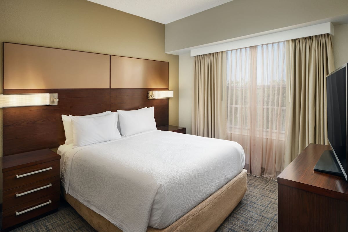 A hotel room with a large bed, a wooden headboard, a bedside table, a TV, and a window with curtains.