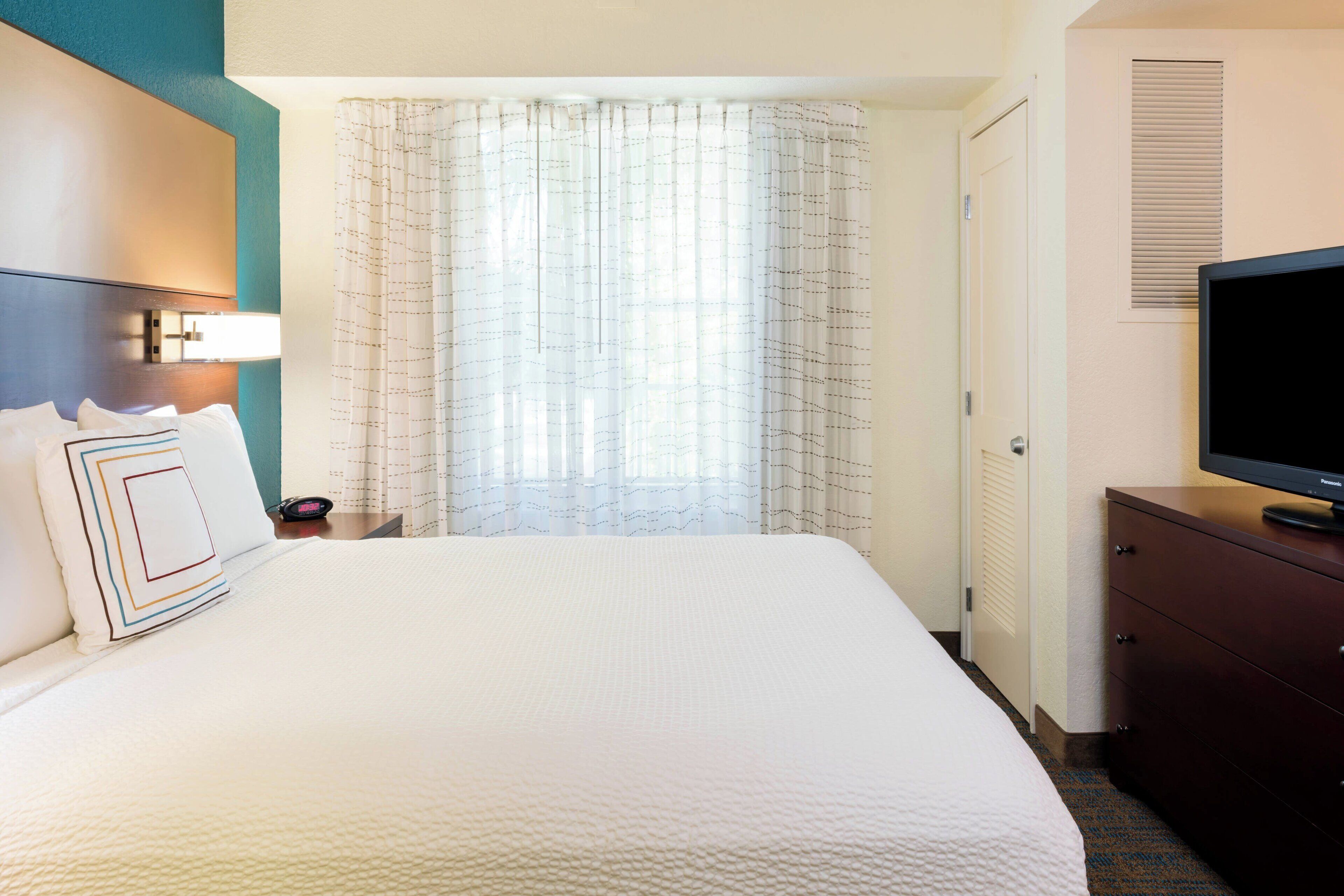 A hotel room with a large bed, a television on a dresser, a nightstand, and a window with curtains.