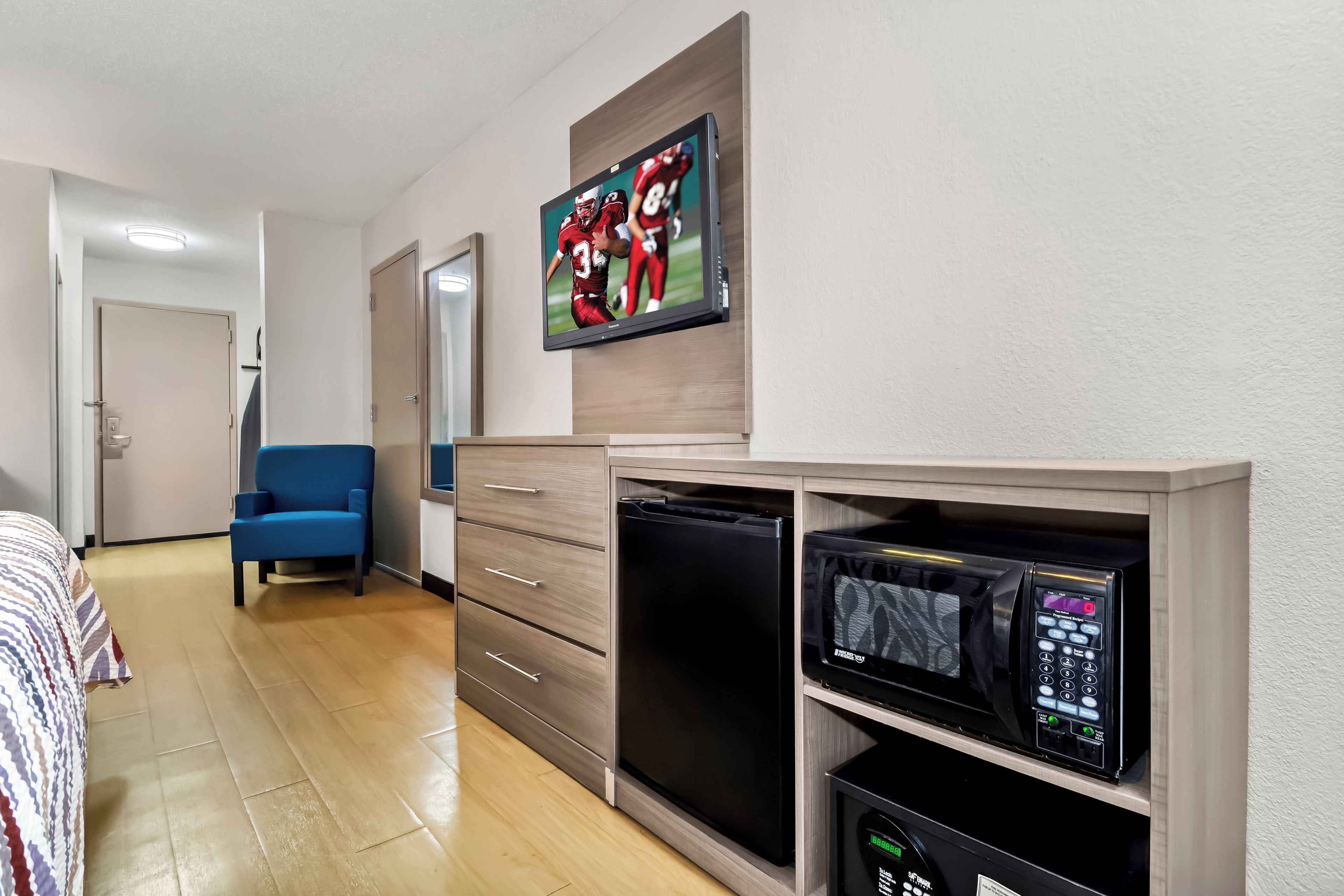A hotel room with a bed, a blue armchair, a television mounted on the wall, and a microwave on a shelf.