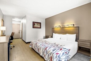 A hotel room with a bed, bedside tables, a desk with a chair, a framed picture on the wall, and a door leading to another room. - Red Roof Inn Myrtle Beach Hotel - Market Common (Myrtle Beach)