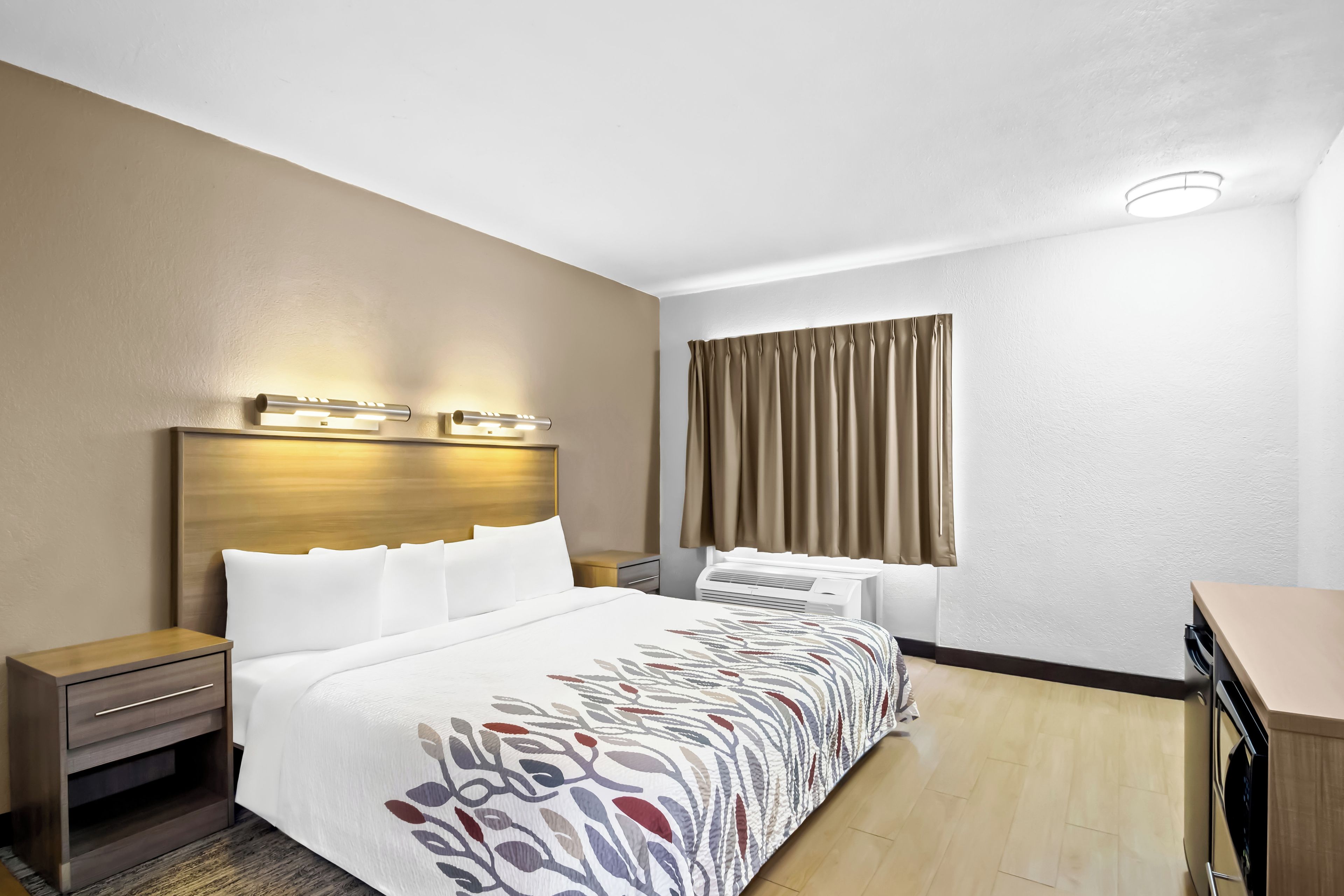 Quarto de hotel com cama, mesas de cabeceira, escrivaninha, janela com cortinas e ar condicionado.