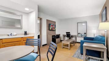 Un salon moderne avec un canapé bleu, une table à manger avec des chaises, un coin cuisine équipé d’un micro-ondes et un téléviseur fixé au mur.
