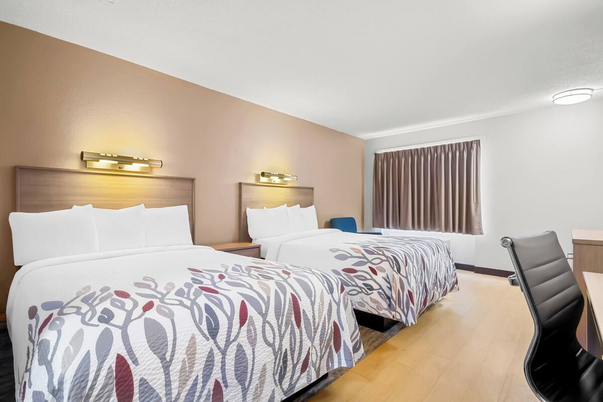 A hotel room with two beds, a desk, a chair, and a window with curtains.